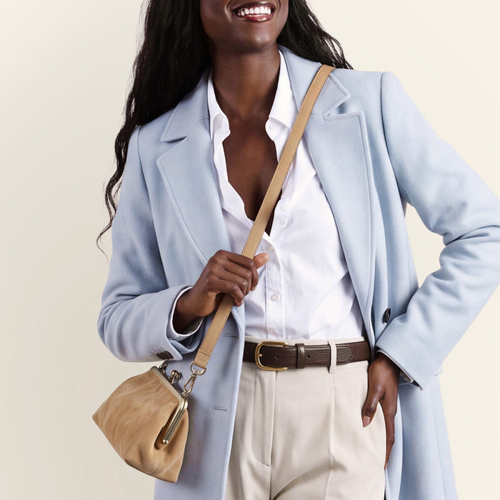 A woman wearing a light blue coat over a white shirt and beige trousers, carrying a beige shoulder bag against a plain cream background.
