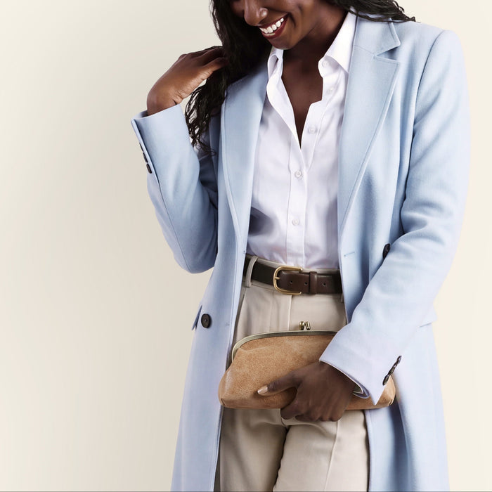 A woman wearing a light blue tailored coat over a white button-up shirt and beige trousers, holding a tan clutch purse against a plain cream background.