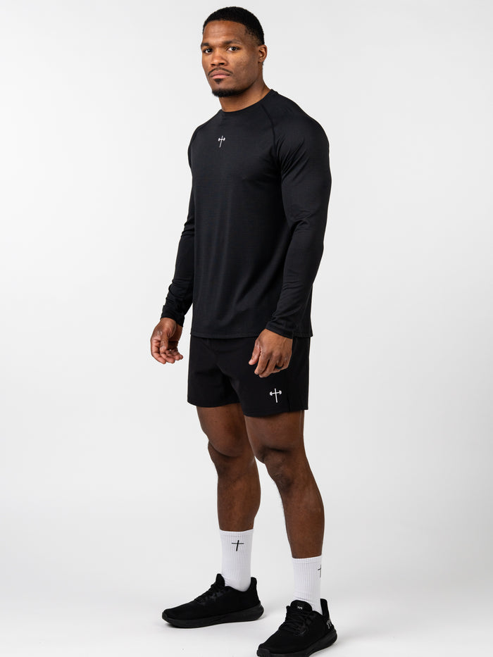 Man wearing the Carry Your Cross Premium Long Sleeve Tee in black with matching black shorts, white socks featuring a cross design, and black athletic shoes, standing against a white background.