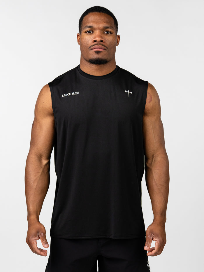A muscular man wearing a black Carry Your Cross Premium Oversized Tank with a small cross and the text "LUKE 9:23" printed on the chest, styled for Christian fitness apparel.