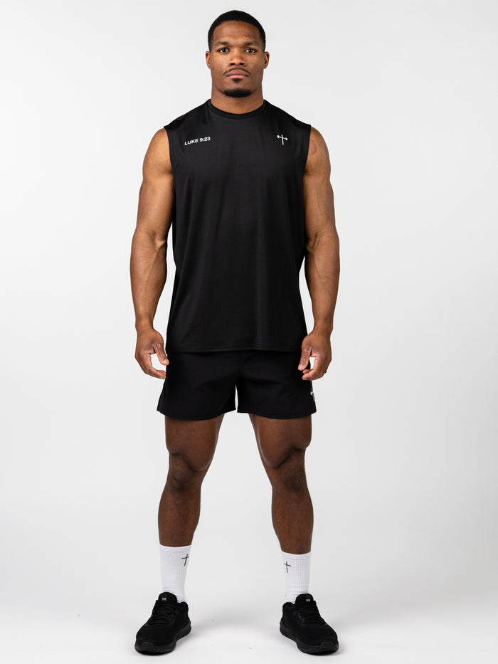 Male model wearing a black Carry Your Cross Premium Oversized Tank with subtle cross and scripture details, paired with black athletic shorts, socks, and shoes.