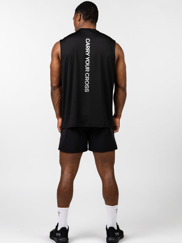 Man wearing the Carry Your Cross Premium Oversized Tank in black, showing the back with vertical "CARRY YOUR CROSS" text, paired with black shorts and white socks with cross detail, representing Christian fitness apparel.