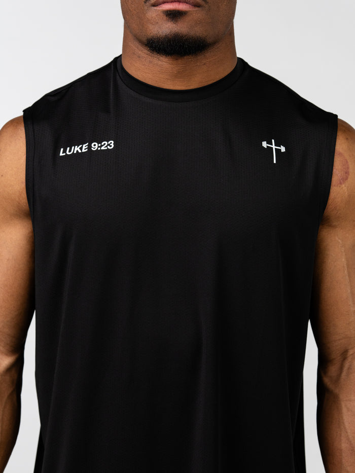 Close-up of a man wearing a black Carry Your Cross Premium Oversized Tank with a small white cross and barbell symbol on the chest and the text LUKE 9:23 on the upper left side, representing Christian gym clothing and faith based fitness apparel.