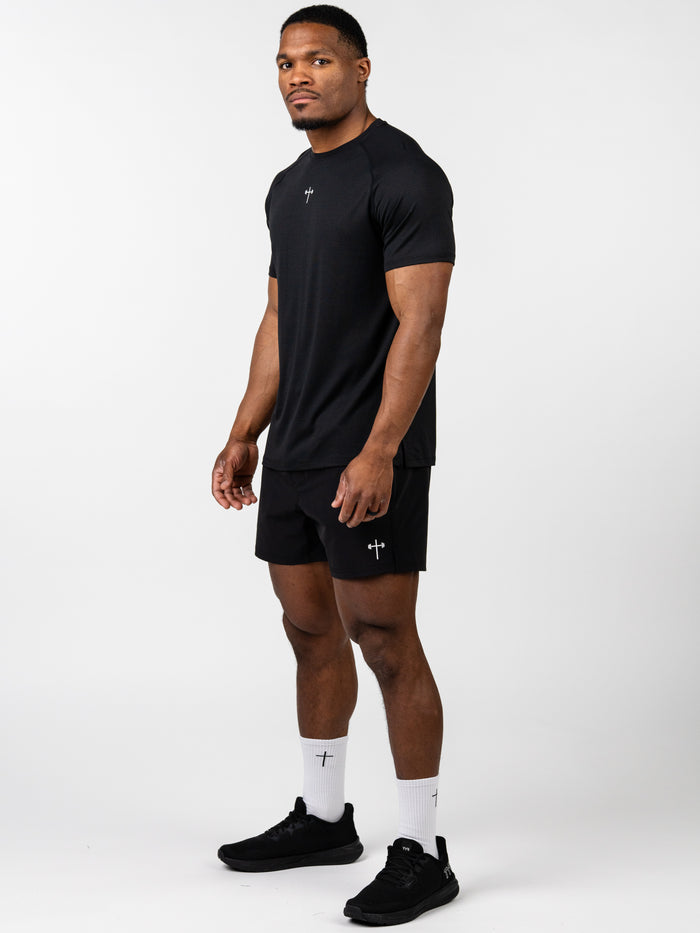 A fit man wearing a black Carry Your Cross Premium Tee with a small cross and barbell logo on the chest, paired with matching black shorts and white socks featuring a cross symbol, posing against a plain white background.