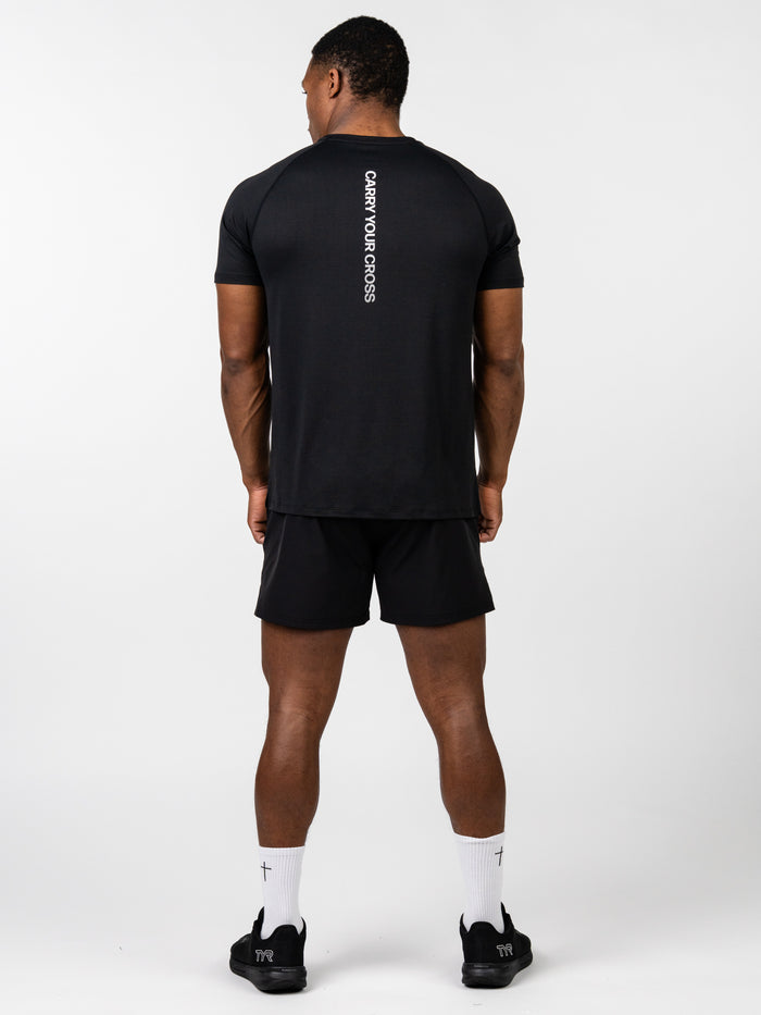 Man wearing black Carry Your Cross Premium Tee with white vertical text on the back, paired with black shorts and white socks with small cross symbols, standing against a plain white background.
