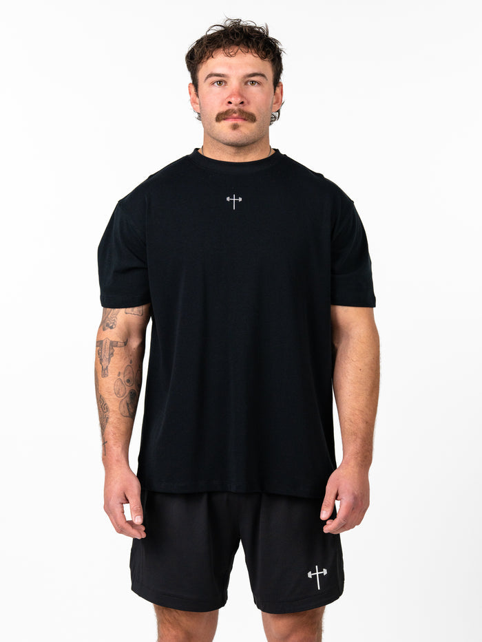 Man wearing a black Christ is King Wing Premium Oversized Tee with a small cross and barbell graphic on the chest, styled with matching black shorts, showcasing Christian fitness apparel.