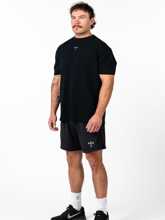 A man wearing the Christ is King Wing Premium Oversized Tee in black, paired with black athletic shorts and white socks featuring cross designs, showcasing faith-based Christian gym clothing.