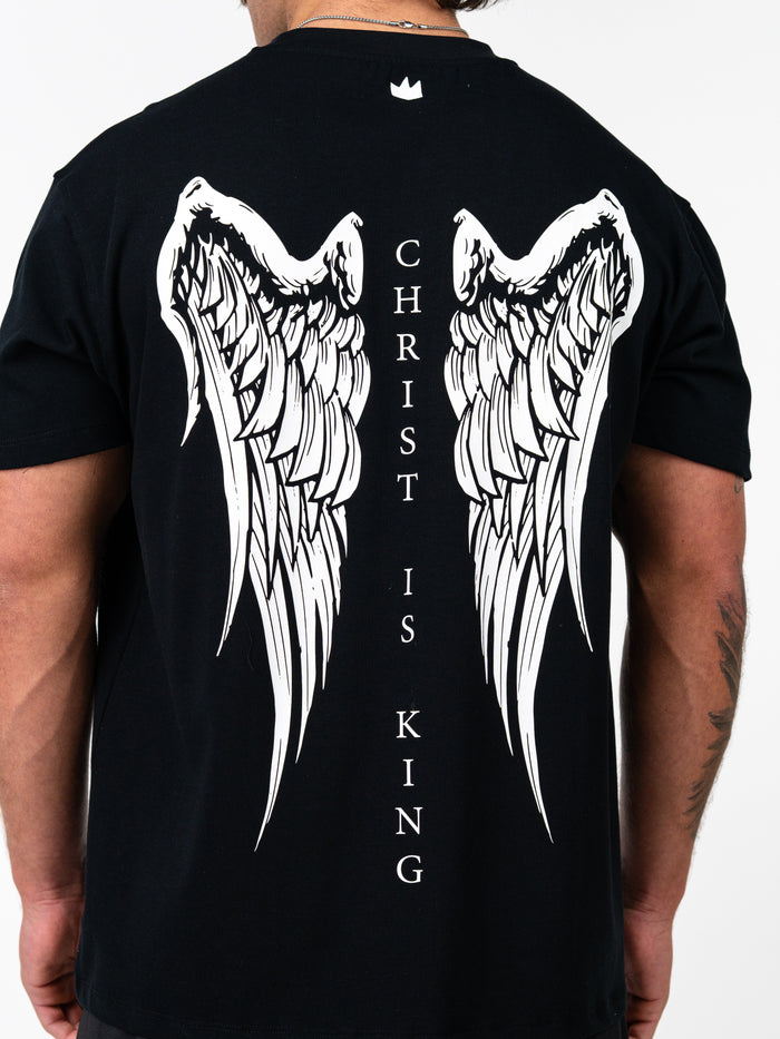 Back view of a person wearing a black Christ is King Wing Premium Oversized Tee featuring large white angel wings and the text "Christ is King" printed vertically between the wings.