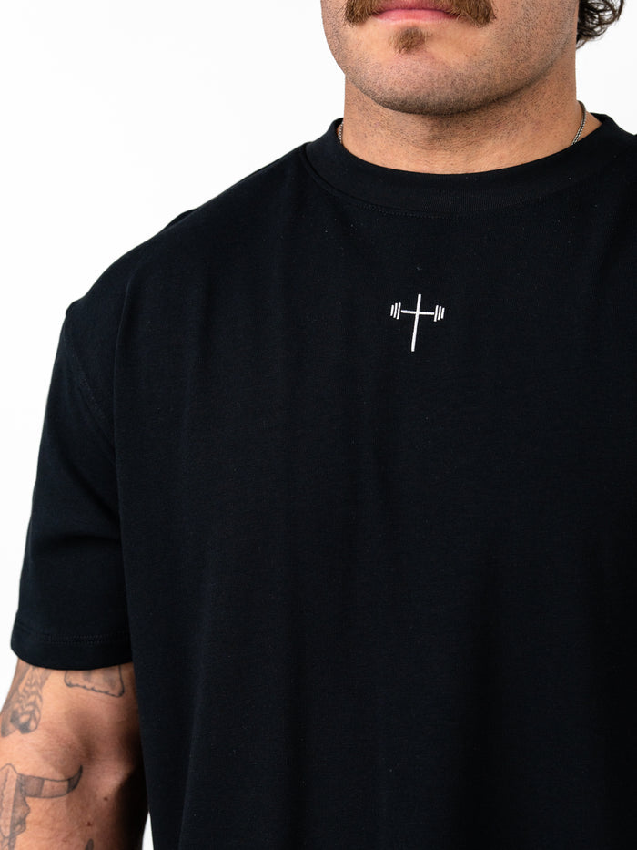 Man wearing a black Christ is King Wing Premium Oversized Tee featuring a small white cross with a barbell design on the chest, showcasing Christian fitness apparel.