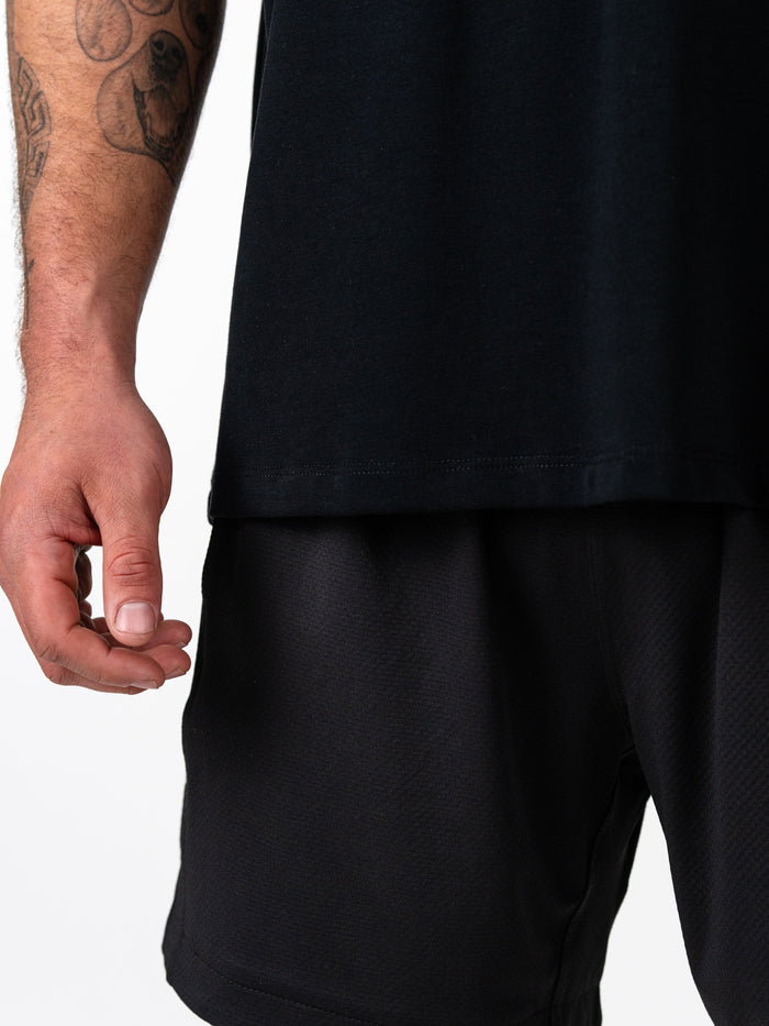 Close-up view of a man's arm with tattoos wearing the Christ is King Wing Premium Oversized Tee in black paired with black athletic shorts, showcasing Christian gym clothing for faith-based fitness.