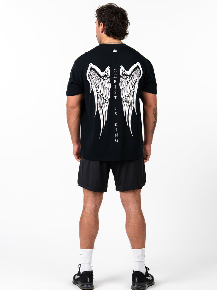 Back view of a man wearing the black Christ is King Wing Premium Oversized Tee featuring large white angel wings and vertical "Christ is King" text, paired with black athletic shorts.