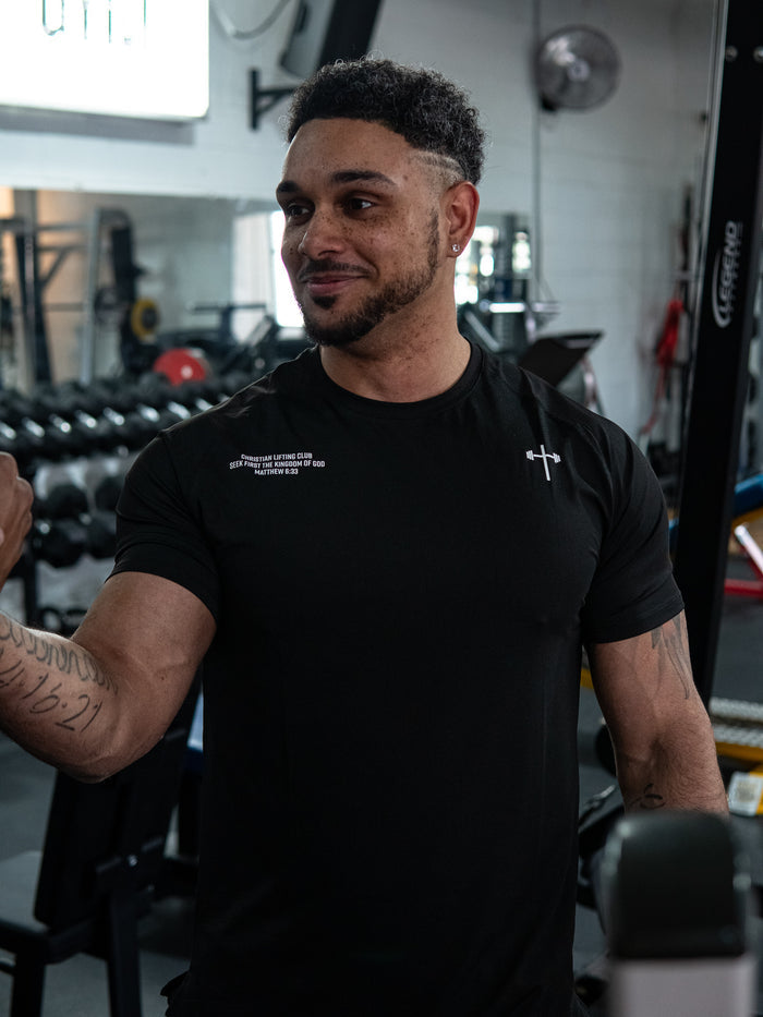 Man wearing a black Christian Lifting Club Premium Fitted Tee with a small cross and barbell logo on the chest, standing in a gym environment with weights in the background.
