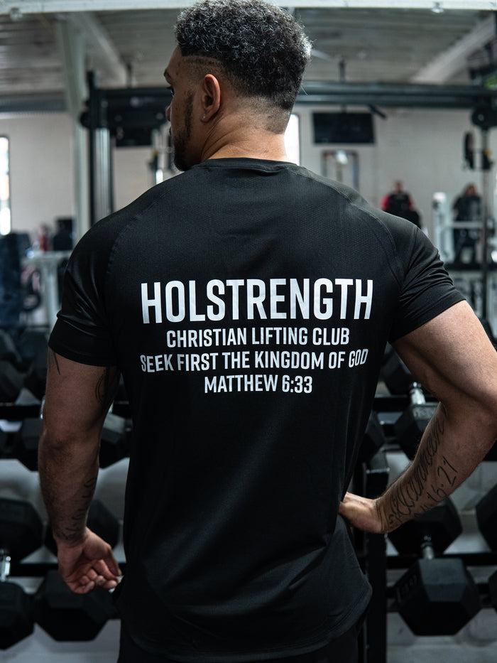 Man wearing a black Christian Lifting Club Premium Fitted Tee in a gym setting, featuring white text on the back with HolStrength branding and a biblical quote from Matthew 6:33.