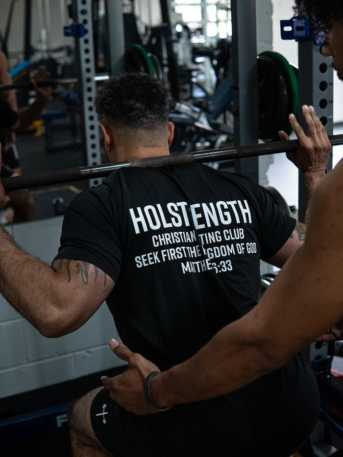 Man lifting weights in a gym wearing a Christian Lifting Club Premium Fitted Tee with faith-based fitness apparel branding on the back.