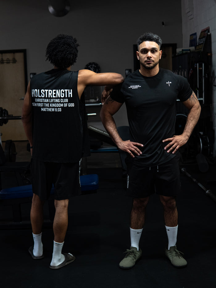Two men wearing Christian Lifting Club Premium Fitted Tee and sleeveless shirt in a gym setting, showcasing Christian fitness apparel with faith-based messages on the clothing.