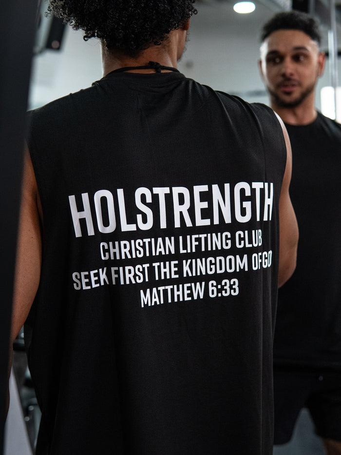 Rear view of a person wearing the Christian Lifting Club Premium Oversized Tank in black with white text that reads "HOLSTRENGTH Christian Lifting Club Seek First the Kingdom of God Matthew 6:33" during a gym workout session.