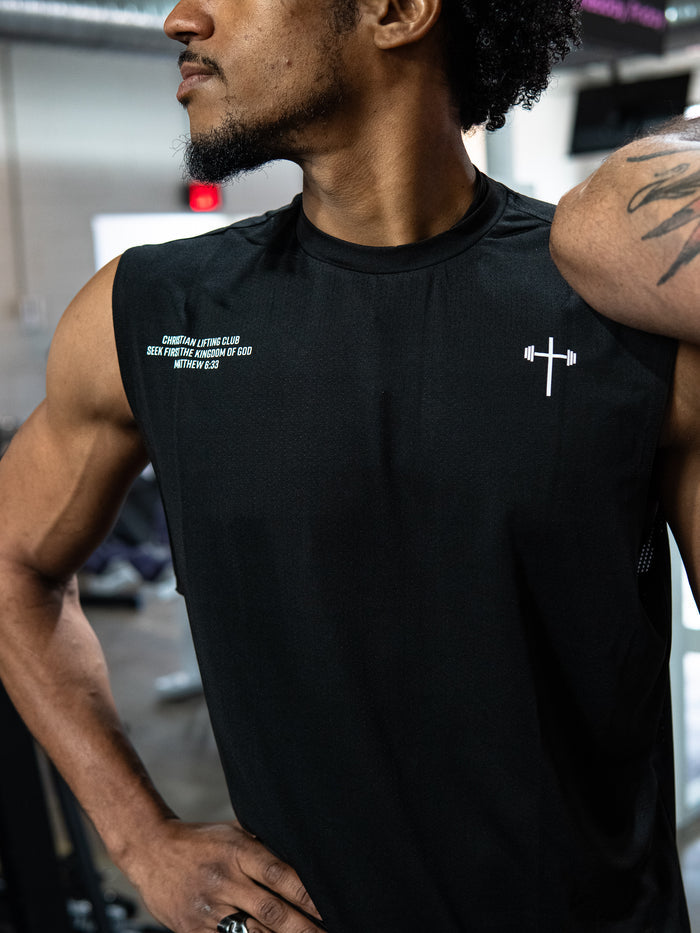 Man wearing a black Christian Lifting Club Premium Oversized Tank with a small white barbell cross logo and inspirational text on the chest, posing in a gym setting.