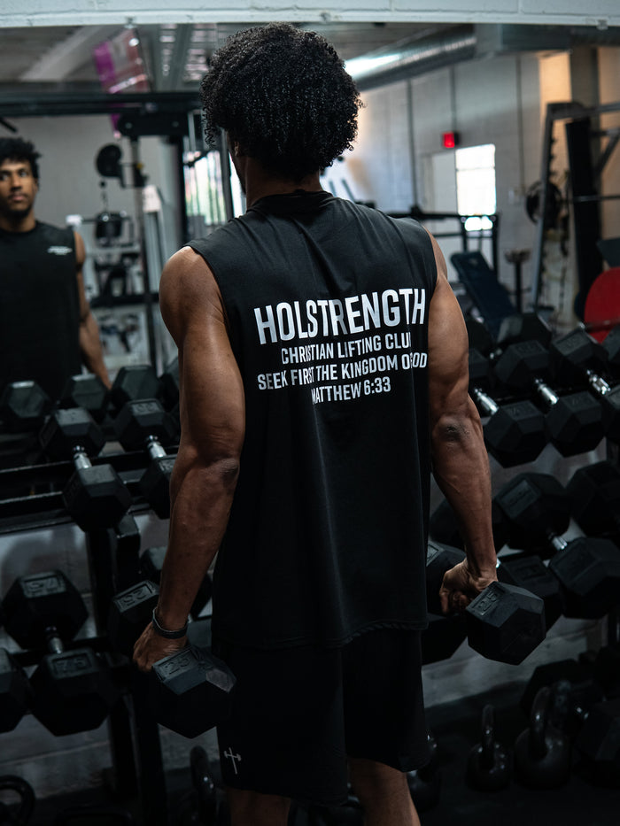 Man wearing a Christian Lifting Club Premium Oversized Tank in a gym, holding dumbbells with back text referencing faith and strength while standing in front of weights and mirror.
