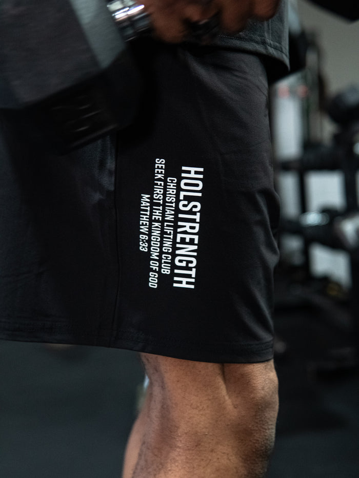 Close-up of a person wearing Christian Lifting Club Premium Shorts 7" black athletic shorts with white text reading "HOLSTRENGTH" and a Bible verse on the side, in a gym setting.