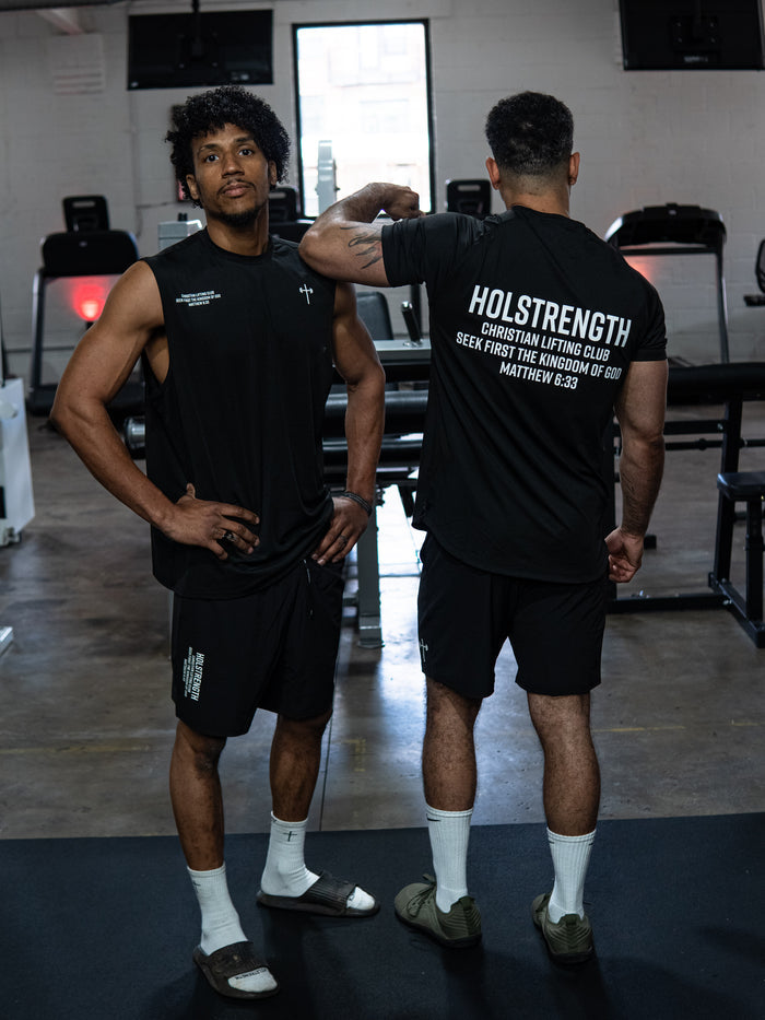 Two men in a gym wearing black Christian Lifting Club Premium Shorts 7" paired with black sleeveless and short-sleeve tops featuring faith-based fitness branding and scripture.