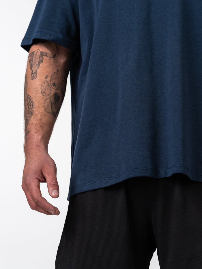 Close-up of a person wearing a navy blue Classic Oversized Tee, showing a relaxed fit and part of a tattooed arm, paired with black athletic shorts.