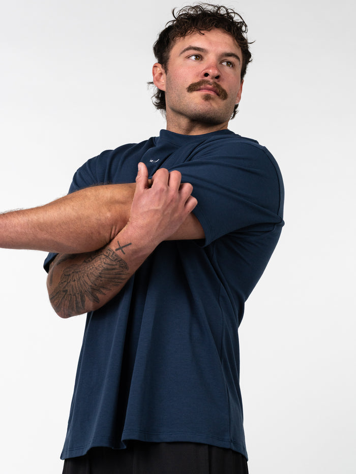 Man stretching arm wearing a navy Classic Oversized Tee, featuring a relaxed fit and designed for movement, styled with visible tattoos on forearm against plain background.