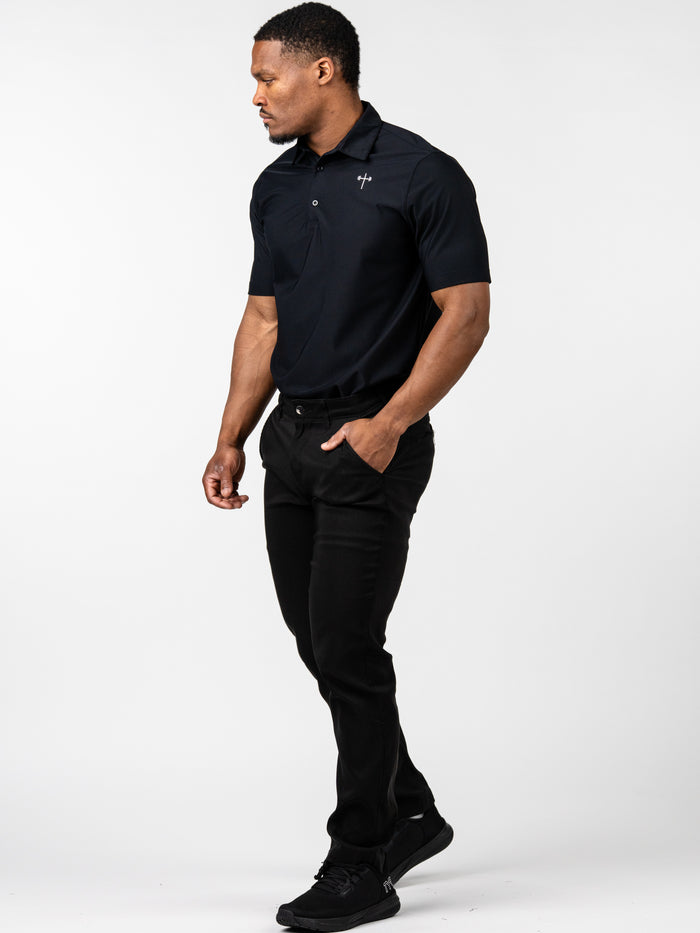 Man wearing a black Core Athletic Polo with a subtle cross and barbell design on the chest, paired with black pants and sneakers, standing against a white background.