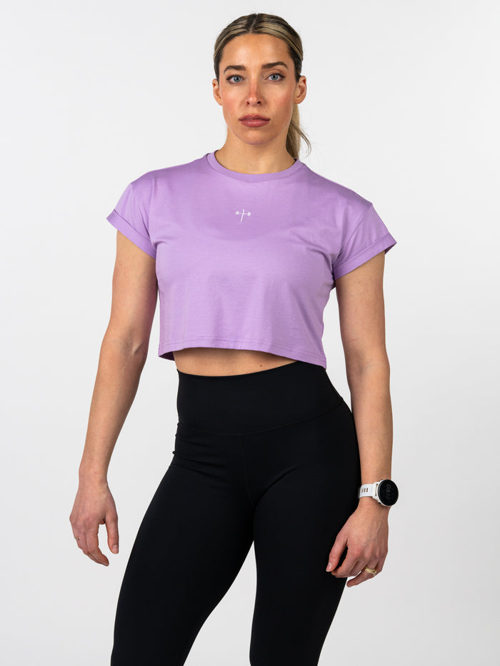Woman wearing a lavender Cross Essential Crop Tee with a small white cross and barbell graphic, paired with black high-waisted leggings, styled for fitness and activewear.