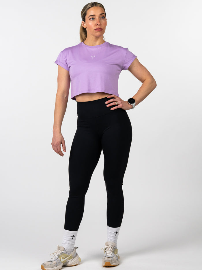 Woman wearing a lavender Cross Essential Crop Tee with a small Christian cross and barbell design on the chest, paired with black leggings, white socks with crosses, and athletic shoes, modeling Christian fitness apparel.