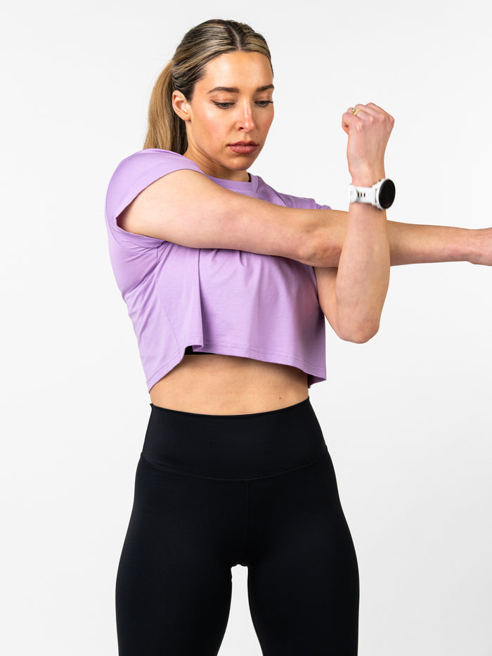 Woman stretching her arm wearing a light purple Cross Essential Crop Tee paired with black high-waisted leggings, demonstrating comfort and performance in Christian activewear.