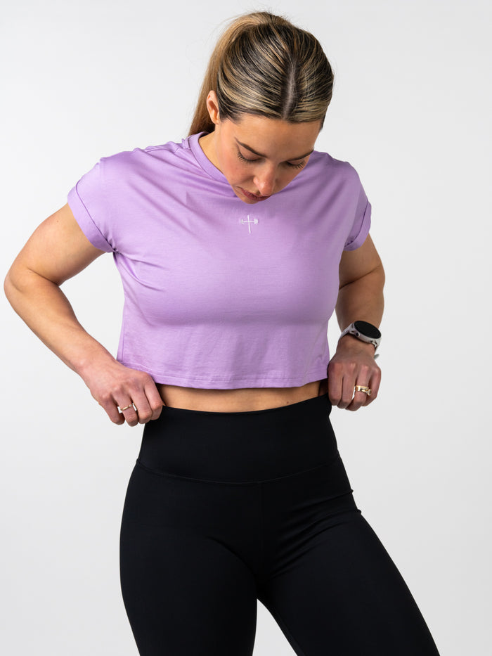 Woman wearing a lavender Cross Essential Crop Tee with a small white cross and barbell design on the chest, paired with black high-waisted leggings, adjusting the hem.