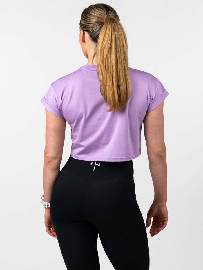 Woman wearing a lavender Cross Essential Crop Tee paired with black leggings that feature a small barbell cross logo on the waistband, showcasing Christian fitness apparel.