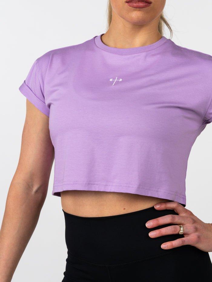 Woman wearing a lavender Cross Essential Crop Tee with a small white cross and barbell design on the chest, paired with black high-waisted leggings in a studio setting.