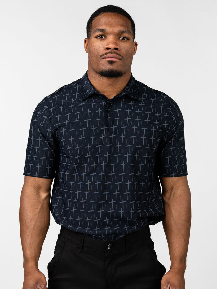 Man wearing a black Cross Premium Polo shirt patterned with subtle gray crosses, paired with black pants, standing against a plain white background.