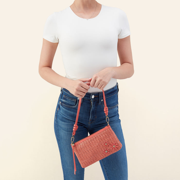 A model wearing a white t-shirt and blue jeans holds a coral pink woven crossbody clutch bag with a matching strap against a light beige background.