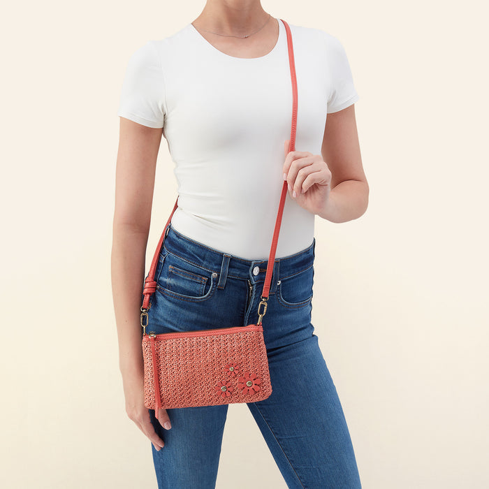 A woman wearing a white t-shirt and blue jeans models a coral pink woven crossbody bag with a matching strap against a solid light beige background.