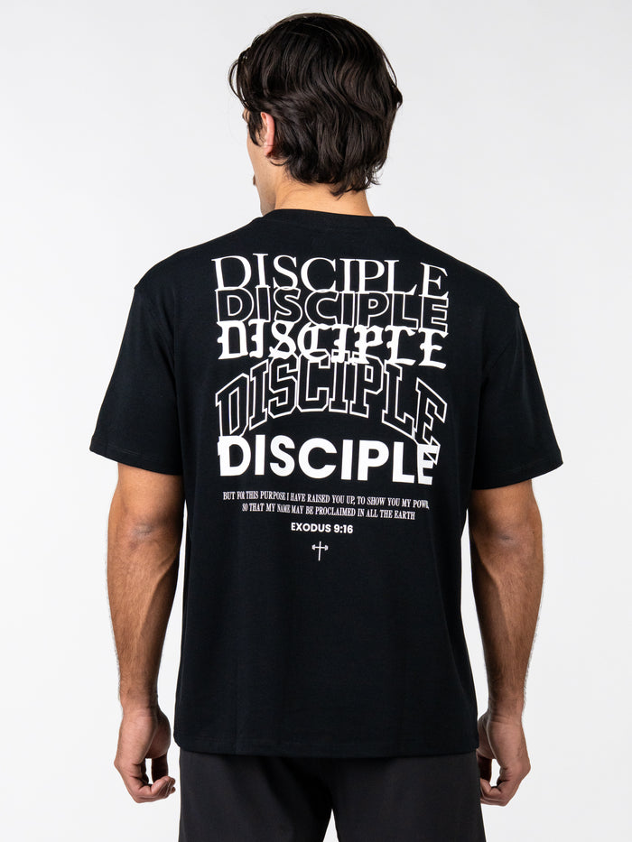 Man wearing a black Disciple Premium Oversized Tee with multiple stylized white "Disciple" text prints and a scripture reference Exodus 9:16 on the back, featuring Christian fitness apparel design.