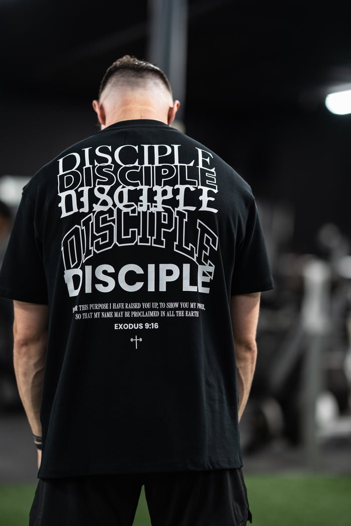 Man wearing a black Disciple Premium Oversized Tee with bold white "DISCIPLE" text and Exodus 9:16 scripture printed on the back, standing in a gym setting.