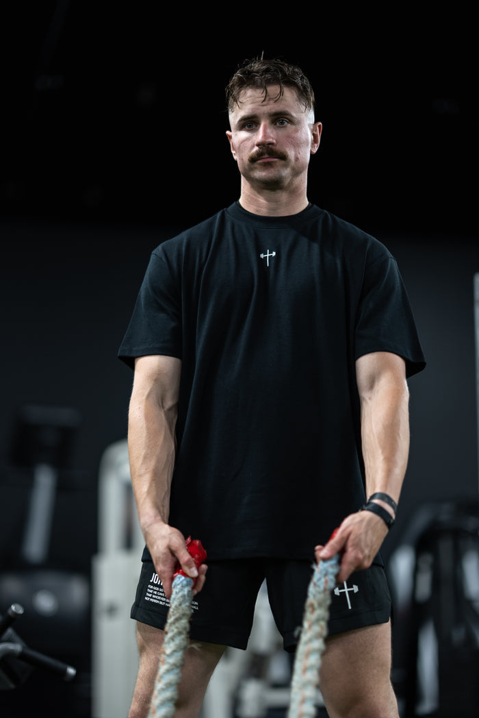Man wearing Disciple Premium Oversized Tee in a gym holding battle ropes, showcasing Christian gym clothing with a minimalist cross design on the shirt.