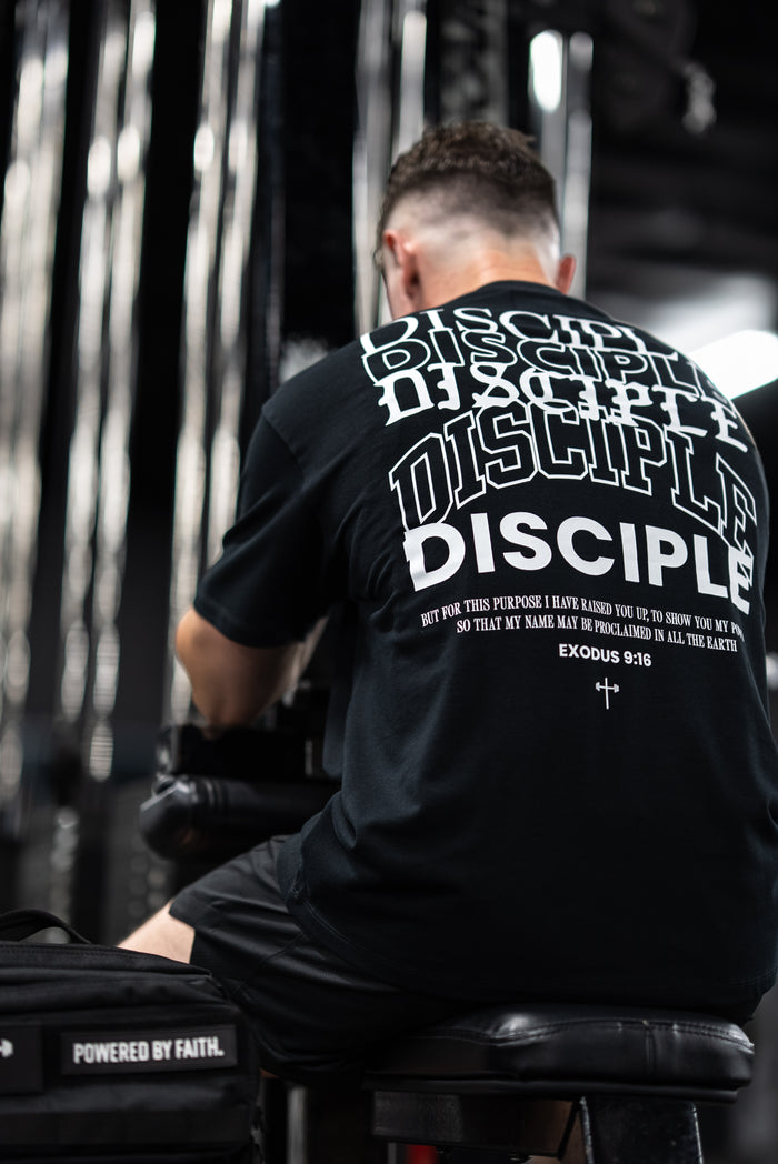 Man wearing a black Disciple Premium Oversized Tee featuring bold repeating text and a Bible verse from Exodus 9:16, sitting in a gym setting with fitness equipment.