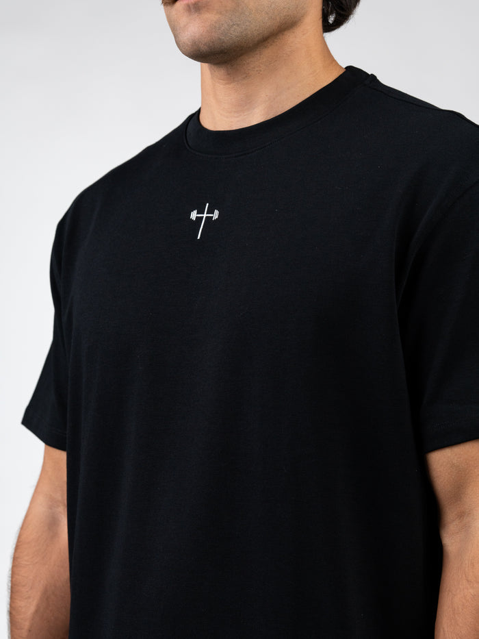 Close-up of a man wearing a black Disciple Premium Oversized Tee featuring a small white cross with a barbell design on the chest, blending Christian gym clothing with faith-based fitness apparel.