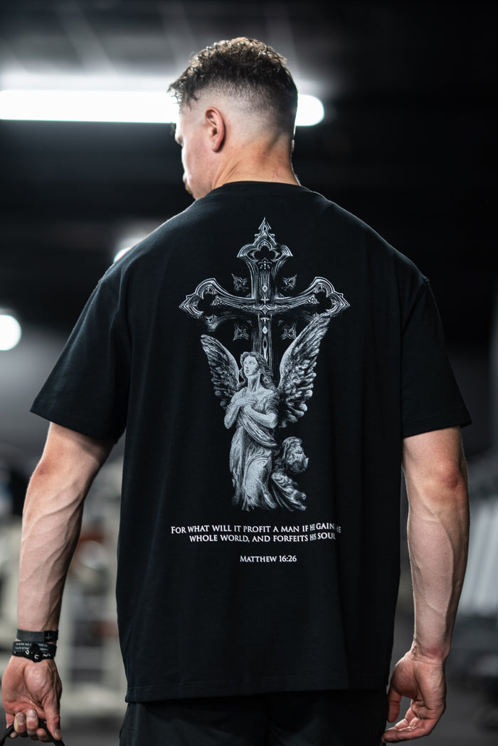Man wearing Divine Angel Premium Oversized Tee featuring a detailed cross and angel graphic with a Bible verse from Matthew 16:26 on the back in an indoor setting.