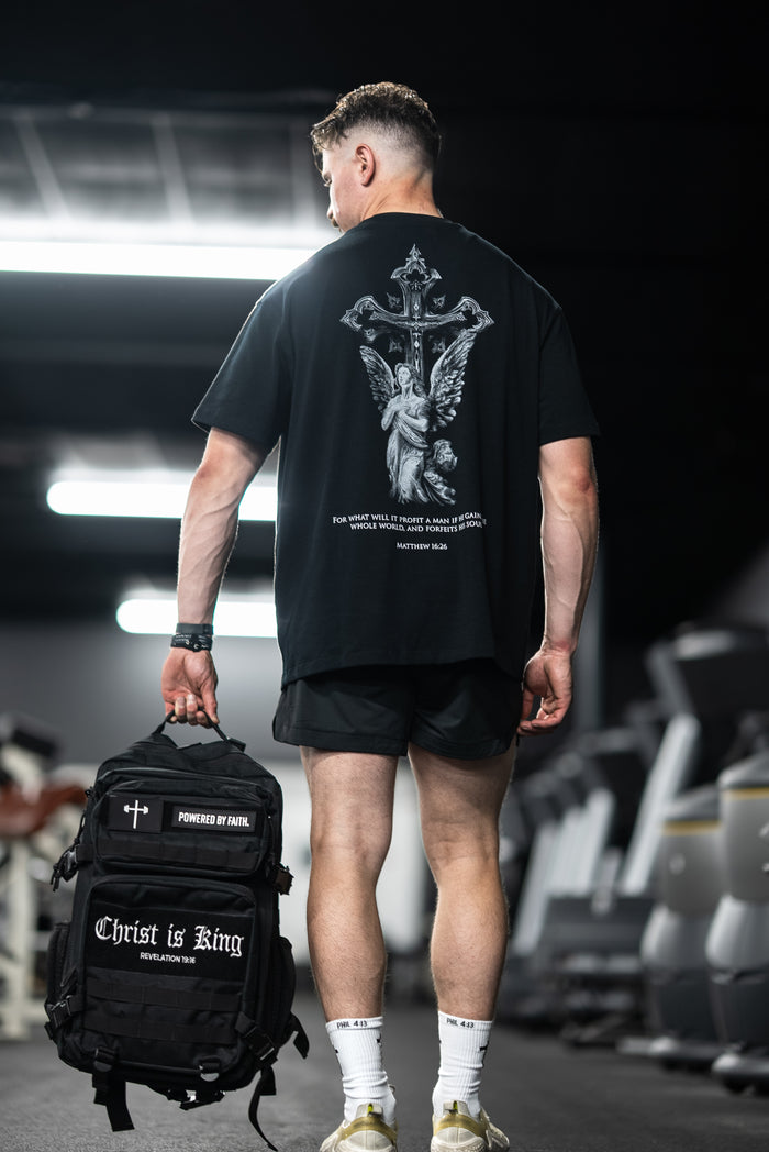 Man wearing a Divine Angel Premium Oversized Tee featuring a detailed angel and cross graphic with a biblical quote on the back, standing in a gym holding a black backpack.