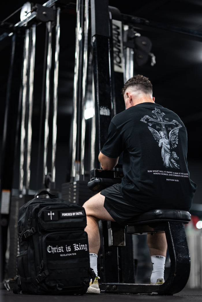 Man wearing Divine Angel Premium Oversized Tee with angel and cross design sitting on gym equipment next to black Christian gym backpack with "Christ is King" text in fitness setting.