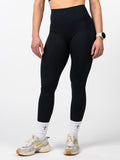 Front view of a person wearing black Essential Leggings paired with white socks featuring a small cross design, athletic shoes, and a fitness watch, highlighting the sleek, squat-proof fit and premium fabric ideal for workouts.