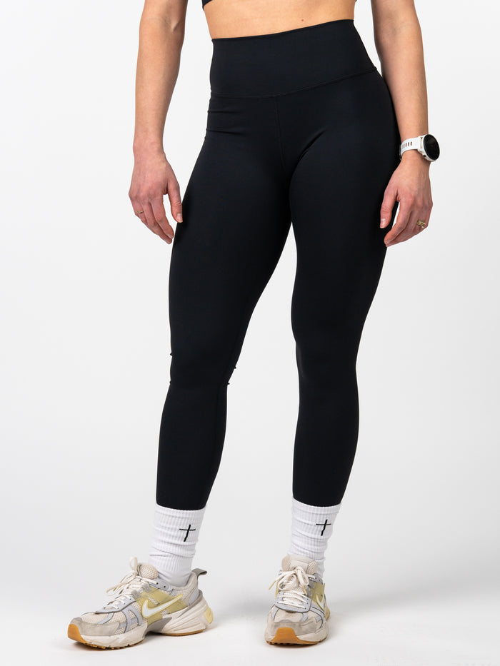 Front view of a person wearing black Essential Leggings paired with white socks featuring a small cross design, athletic shoes, and a fitness watch, highlighting the sleek, squat-proof fit and premium fabric ideal for workouts.