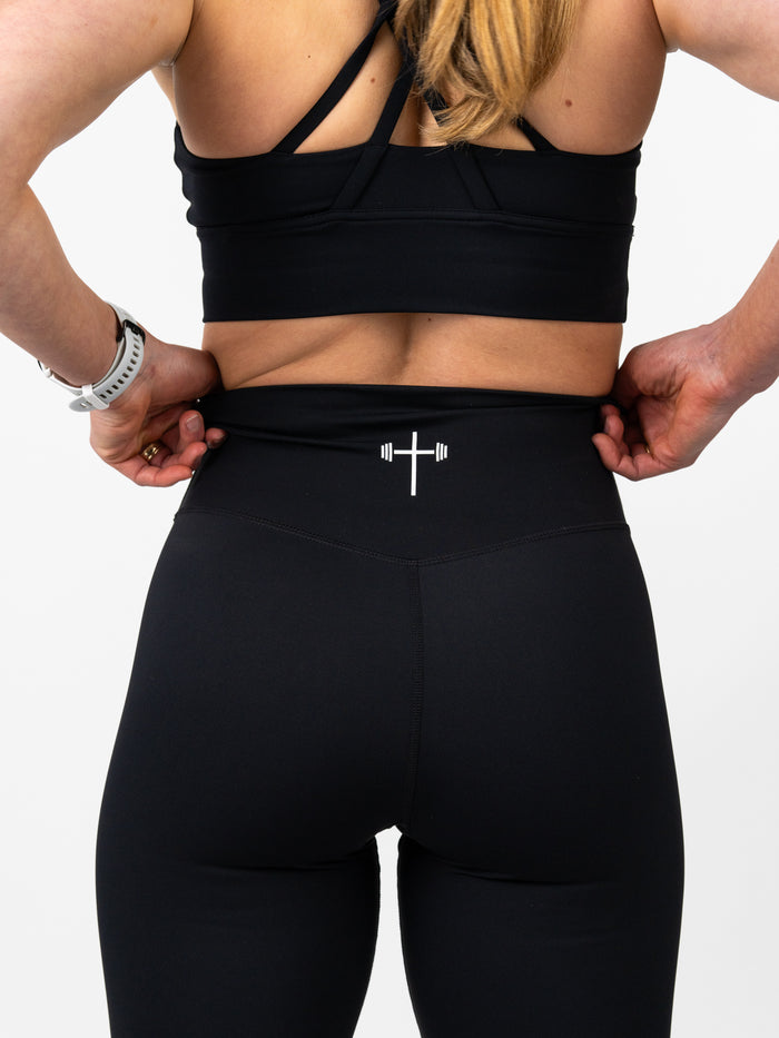 Rear view of a woman wearing black Essential Leggings featuring a subtle Christian cross and barbell design on the waistband, paired with a black sports bra, highlighting faith-based fitness apparel.