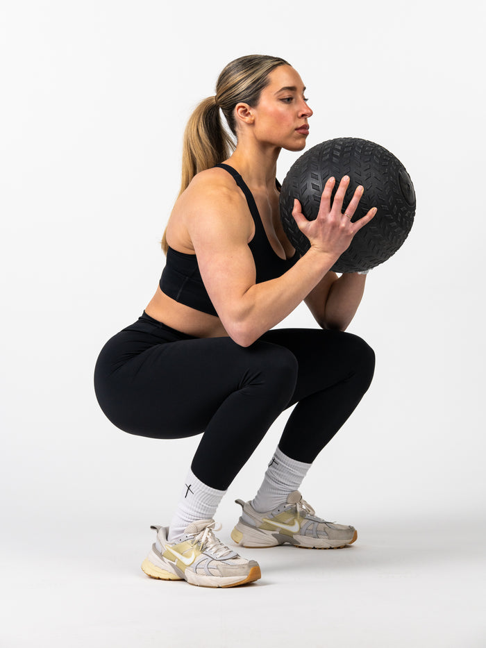 Athletic woman wearing Essential Leggings and a sports bra performing a weighted squat exercise holding a textured black medicine ball against a white background.