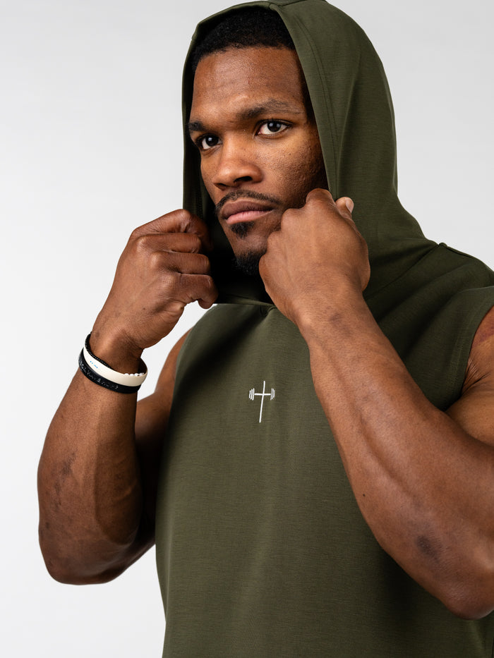 Close-up of a muscular man wearing an olive green Essential Sleeveless Hoodie with a small Christian cross and barbell symbol on the chest, adjusting the hood.