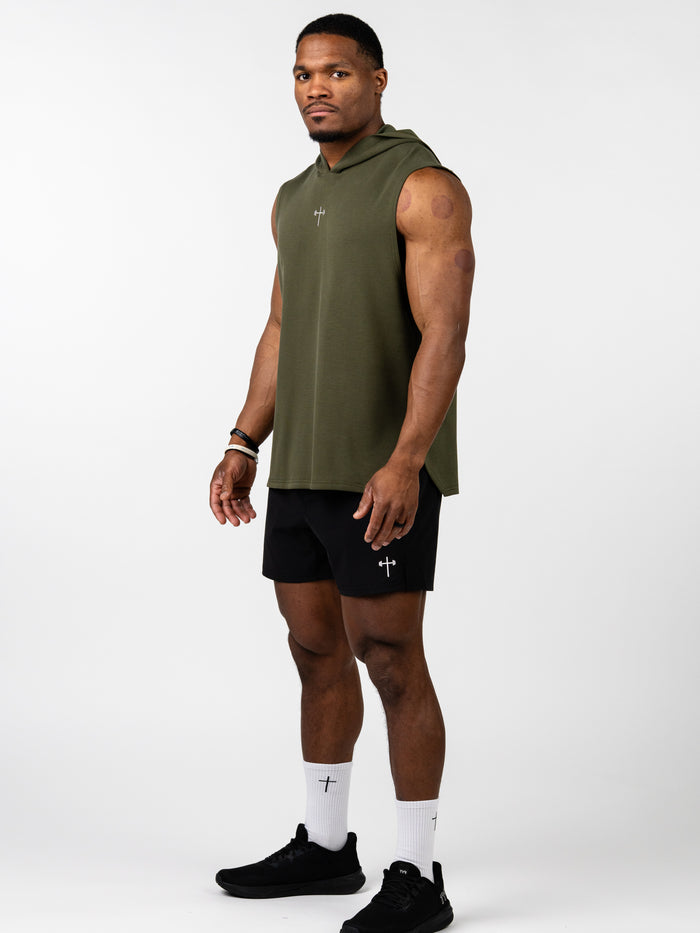 Man wearing an olive green Essential Sleeveless Hoodie with a small cross and barbell design on the chest, paired with black shorts, white socks, and black athletic shoes, modeling Christian fitness apparel.
